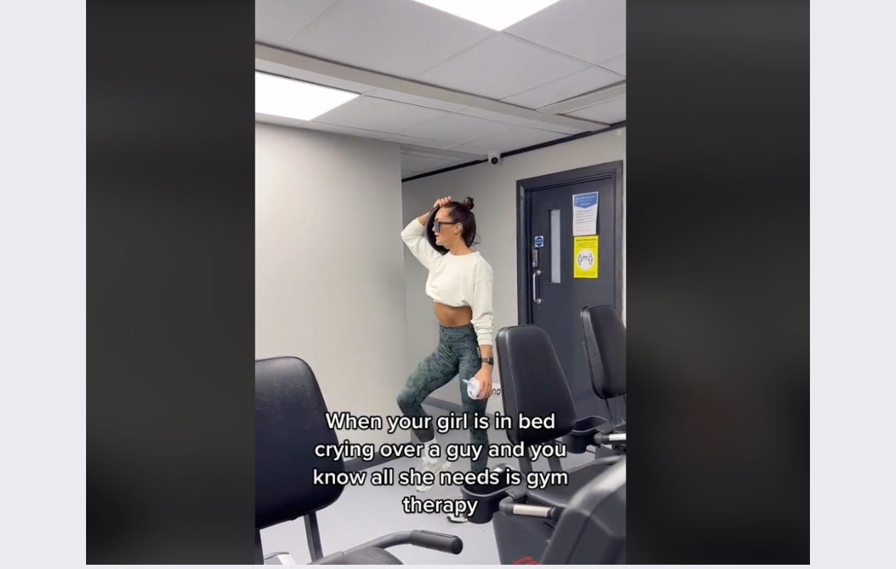 screenshot from gymshark's tiktok video with a girl wearing a white top and grey leggings in a gym, carring a heavy weight